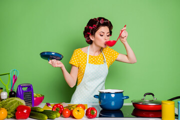 Photo retro stylish girl chef prepare veggie soup boil sauce pan try eat ladle table tomato cucumber wear hair rollers dotted yellow dress skirt isolated green color background