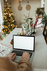 Young woman work on laptop computer with blank display screen with mockup copy space. Cozy comfortable kitchen with table, Christmas tree, fir branches, cones, candles. Holidays celebration.