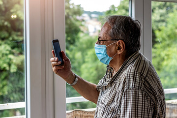 Senior man using mobile for connection with his family during coronavirus isolation. Elderly man with face mask using smart phone indoor. Close up of a senior man consulting with a doctor on his phone