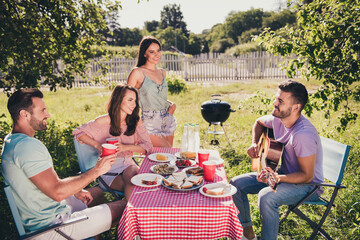 Photo of four people best fellows have summer vacation feast brown hair man musician player play guitar song sit table his friends listen hold drink punch cup outdoors garden