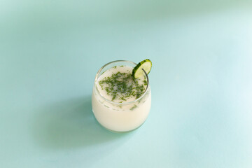 Obraz na płótnie Canvas Ayran in a glass glass with dill and a slice of cucumber on a blue background. Fermented product concept. Copy space.