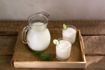 Two glasses with Ayran with a cucumber and a glass jug on a wooden tray on a wooden background. Fermented product concept. Horizontal orientation.