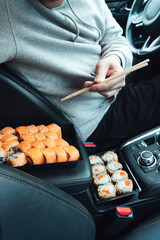 Man is eating sushi, Philadelphia rolls with fish, rice, salmon, cream cheese, avocado in car. Fast, safe, takeaway food on the road to coronavirus pandemic. Delicious japanese snacks with chopsticks