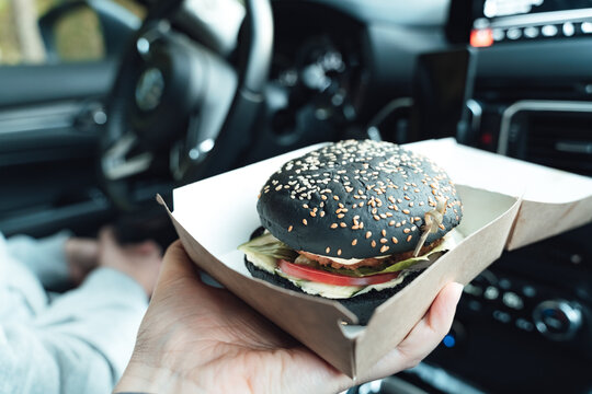 Man is eating black burger with meat, fish, tomato, salad, sause in car. Fast, safe, takeaway food on the road to coronavirus pandemic. Delicious american snack. Family dinner in trip, travel