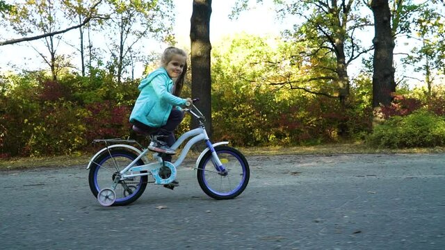 Little girl riding four wheeled bicycle for children along road, trees on background. Dolly shot of happy kid biking in park on sunny day. Concept of sport