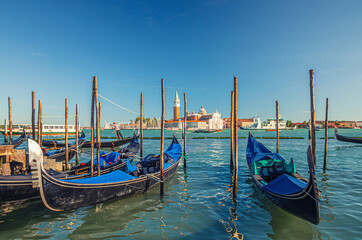 Fototapeta na wymiar Gondolas moored docked on water in Venice. Gondoliers sailing San Marco basin waterway. San Giorgio Maggiore island with Campanile San Giorgio in Venetian Lagoon, blue clear sky, Veneto Region, Italy