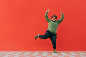 Funny fat guy dancing on a red wall and listening to music on headphones. Cheerful overweight man and casual clothes shows a dance performance.