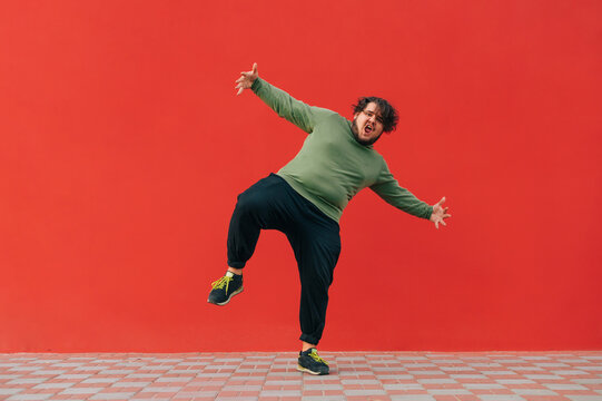 Сharismatic fat man in headphones shows a dance performance on the street against the backdrop of a red wall. Fat guy with curly hair dances on a red background.