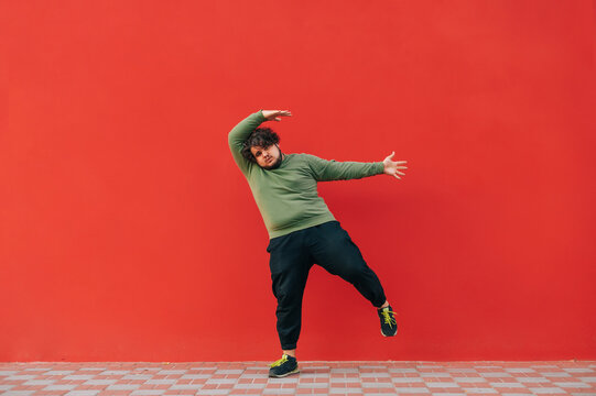 Funny young man with overweight and curly hair dances against the background of a red wall, looks at the camera and moves.