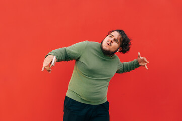 Charismatic fat man with curly hair in casual clothes dancing on a red background and listening to...