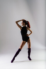 Young beautiful flexible female in black jumpsuit and high heels is posing in a dance studio.