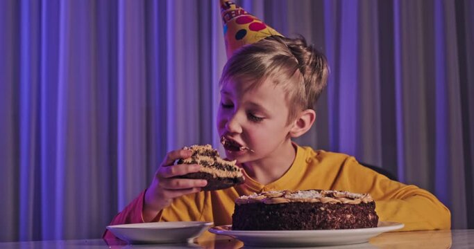Happy child takes a piece of celebration cake and then bite it and swallow.  Cute boy eats cake on his birthday. Happy boy with cone on head celebrates his birthday. Caucasian child and birthday cake