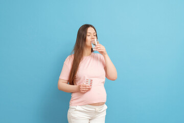 young beautiful pregnant woman on a blue background holds pills in her hand and a glass of water, woman drinking pills