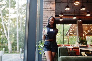 Obraz premium Fashionable feminist african american woman wear in black t-shirt and shorts, posed at restaurant with lemonade glass.