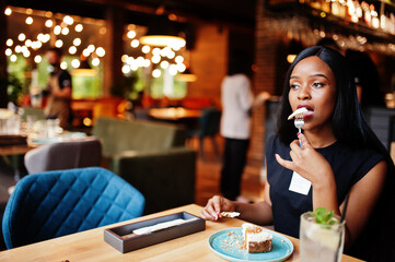 Fashionable feminist african american woman wear in black t-shirt and shorts, posed at restaurant, eat cheese cake.