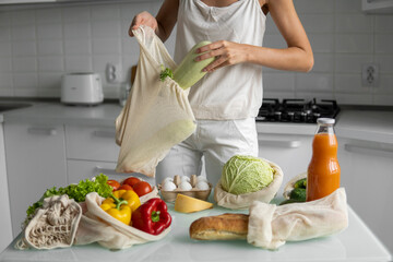 Woman's hand, holding a reusable grocery bag with vegetables on a kitchen at home and takes zucchini out. Zero waste and plastic free concept. Mesh cotton shopper with vegetables. Ecology.