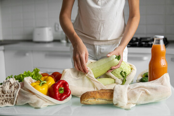 Woman's hand, holding a reusable grocery bag with vegetables on a kitchen at home and takes zucchini out. Zero waste and plastic free concept. Mesh cotton shopper with vegetables. Ecology.