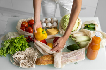 Woman's hand, holding a reusable grocery bag with vegetables on a kitchen at home and takes pepper out. Zero waste and plastic free concept. Mesh cotton shopper with vegetables. Ecology.