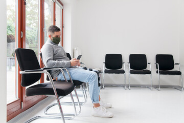 Young man with face mask sitting in a waiting room of a hospital or office looking at smartphone