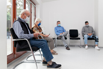 Senior couple with face masks sitting in a waiting room of a hospital together with a young and...