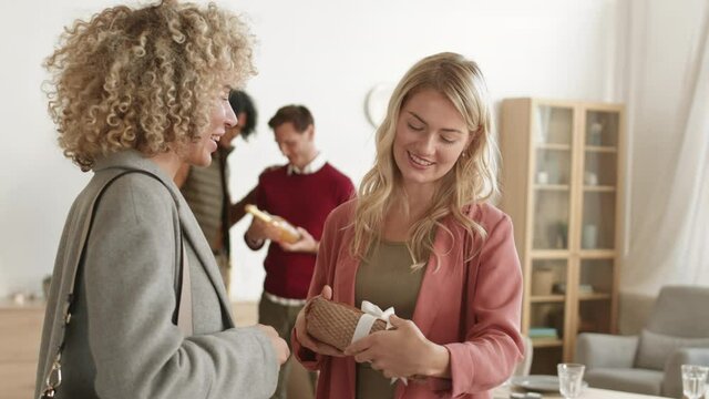 Medium shot of Caucasian curly-haired woman giving gift to woman with blond hair while two diverse men discussing wine bottle on background