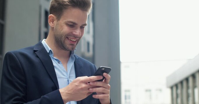 Close Up Portrait of Handsome Successful Businessman is smiling Charmingly while browsing Internet. Happy Caucasian Man is using Smartphone outdoor in Business District, Wearing stylish Suit.