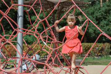 cute little three year old child in a red dress in a rope spider web at the playground