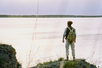 Rear view of young male backpacker in hat standing on edge of cliff and contemplating lake while enjoying hike