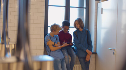 Happy diverse teenage children sitting on sill in school lavatory and play on smartphone