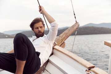 handsome man with a beard at sunset on a yacht wearing a white linen shirt and pants looking at the sea