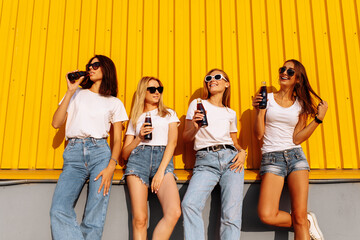 Fototapeta premium happy girlfriends posing and having fun and drinking drinks on the background of the yellow wall of the shopping mall on a sunny summer summer day