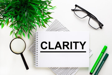 On a white background lies a notebook with the word CLARITY, glasses, a magnifying glass, green markers and a green plant
