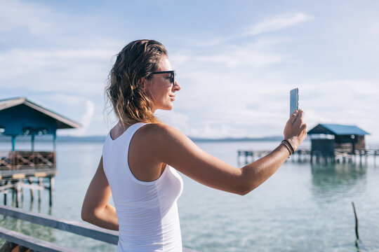 Attractive Caucasian wanderer using cellphone gadget for shooting selfie video and share influence stream content to social networks, millennial tourist clicking pictures during resort vacations