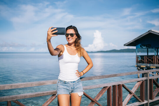 Cheerful Caucasian girl in stylish sunglasses using mobile and making selfie pictures and share positive content to blog followers, happy female tourist enjoying summer vacations for visiting Maldive