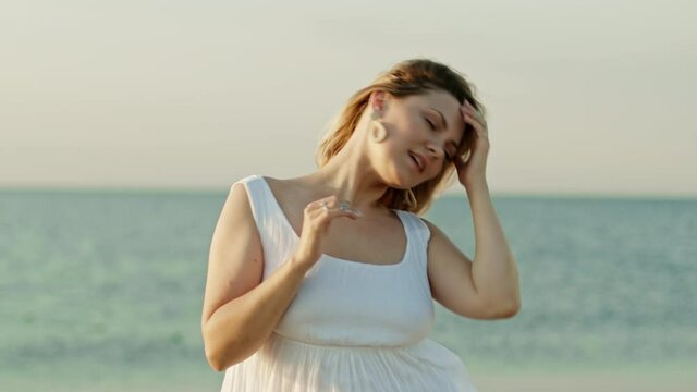 Woman in white dress with blonde hairstyle dancing head on sea background. Girl moves to rhythm of music. Female having fun. She smiling, hair flutter beautifully. Amazing positive footage.