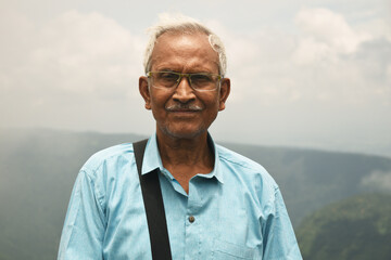 An old man, wearing spectacles and blue shirt, standing alone in nature a blurry mountain background. The elder person with a side bag on his shoulder, looking towards the camera. Copy Space For Text.