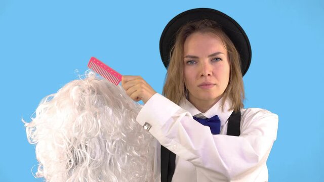 Woman Combing White Wig With Pink Comb on Blue Background in Studio, Looking Serious Look. Stylish hairdresser dressed in a white shirt, bow tie and hat.