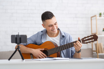 Young student learning to play guitar during self-isolating at home or online tutor. Smiling guy looks at guitar and makes video lesson for followers in living room during virus outbreak