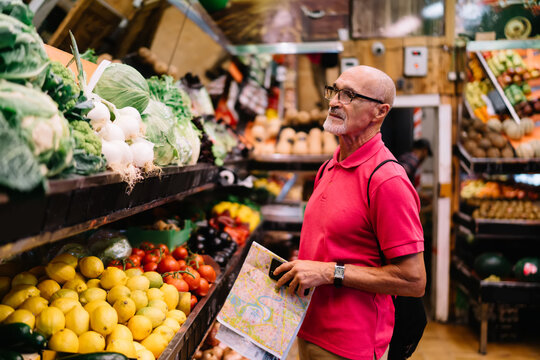 Mature Caucasian senior in optical eyewear for vision correction holding orientation location map in hand and looking for fresh vitamins during shopping, aged gourmet enjoying healthy lifestyle