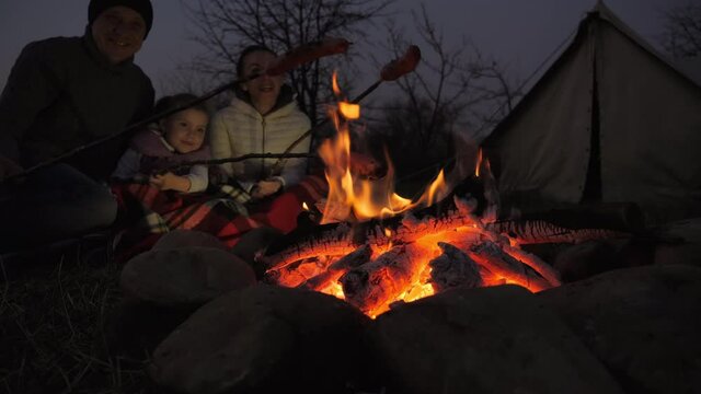 Parents And Their Little Daughter Fry Sausages On The Fire Near A Tourist Tent. The Family Sits Around A Campfire On A Summer Late Evening Weekend In Nature, Cheerful Company, And Happy Faces.