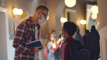 Side view of young teacher talking to african schoolboy wearing safety mask in school corridor