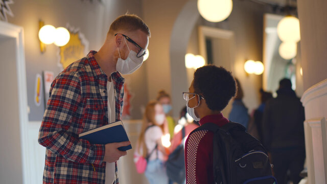 Side View Of Young Teacher Talking To African Schoolboy Wearing Safety Mask In School Corridor