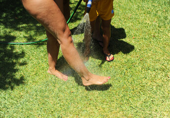 Hijo bañando con el agua de la manguera las piernas de su madre en el jardín de la casa de la playa