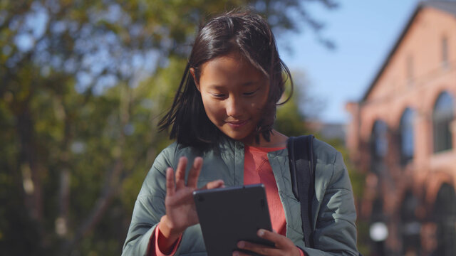 Happy asian girl student with backpack using digital tablet standing outdoors school building