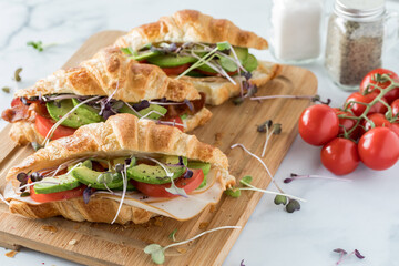 Three croissant sandwiches on a wooden board surrounded by tomatoes on the vine and salt and pepper shakers.