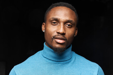 dark-skinned handsome man in a blue jturtleneck on a dark background looks at camera