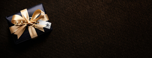 A Christmas present wrapped in black paper and gold ribbon with a blank tag, card against a dark background from above.