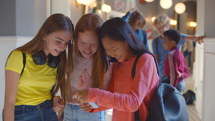 Three multiethnic school girlfriends using smartphone in corridor during break
