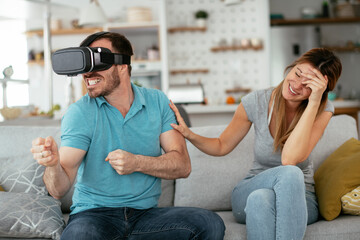 Fototapeta premium Smiling young man using VR headset at home on couch. Woman and her husband enjoying virtual reality in apartment..
