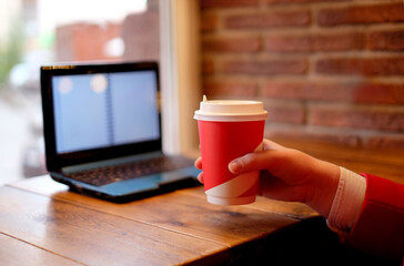 Women hand close up holds red cup on a brick wall background bokeh and open laptop, freelance style